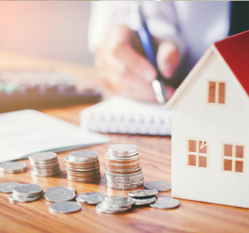 A miniature toy house with coins stacked by the side and a man writing on a notepad